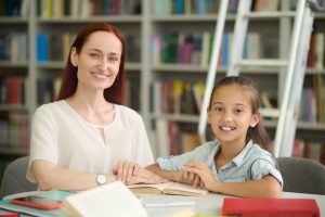 Good mood. Smiling young long-haired woman and primary school girl in casual light clothing doing lessons at table in library looking at camera